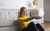 A blonde woman, wearing a yellow turtleneck and jeans, sits on the ground, leaning on a white sofa, while clicking a TV remote control