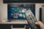 An in-focus hand holds and points a black TV remote at a living room TV blurred in the background.