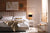 Gray upholstered bed with white and beige linens against a natural wood wall. To the right of the bed is a white nightstand with a black table lamp and sunlight streams through a window.