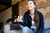Young woman holding a TV remote while sitting on a couch in front of a brick wall. The TV remote is in focus while she and the TV background are out of focus.